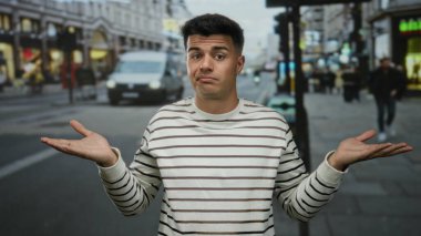 Çizgili gömlekli genç adam hareketli bir şehir caddesinde duruyor elleri havada ve bulanık bir trafik geçmişinin önünde omuz silkiyor..