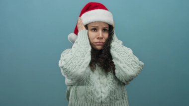 Noel Baba şapkası ve süveteri giyen kadın, elleri başının üzerinde, izole edilmiş mavi bir arka plana karşı hayal kırıklığına uğramış gibi görünüyor..