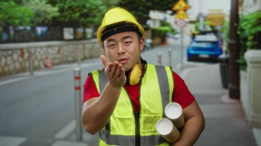 Şehir caddesinde gülümseyen planlı genç bir adam, sarı bir şapka ve yansıtıcı bir yelek giyiyor, kentsel yapıyı somutlaştırıyor..