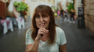 İspanyol kadın şehir arka planında bulanık bir sokakta sessiz hareketler yapıyor, canlı şehir yaşamını sergiliyor ve dinamik bir açık hava ortamında sessizliğini ifade ediyor..