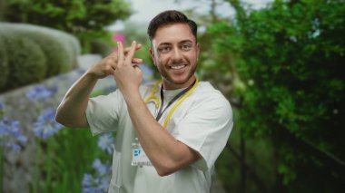 Sakallı ve steteskoplu genç bir erkek doktor parkta gülümsüyor, yeşil ve çiçeklerle çevrili, parmaklarıyla bir etiket işareti yapıyor..