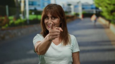Şehir dışındaki bir sokakta enerjik bir şekilde gülümseyen ve işaret eden bir kadın güneşli gökyüzünün altında canlı ve çekici bir ifade yakalıyor..