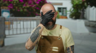 Sakallı bir adam açık havada, şehir ortamını işaret ederek gülümsüyor odak çiçekli arka planda önlük dövmeleri takıyor ve olay yerine renk ve enerji katıyor.