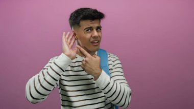 Genç adam pembe arka planda kafa karışıklığı gösteriyor, çizgili kazak ve sırt çantası giyiyor, merakını dile getiriyor ve dinliyor..