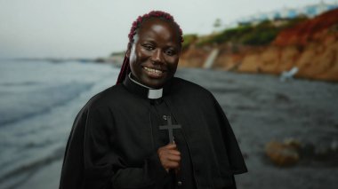 Arka planda haç kolyeli, okyanus dalgaları ve uçurumlarla deniz kenarında gülümseyen kadın, dingin doğal ortamda neşe ve maneviyat yayıyor..