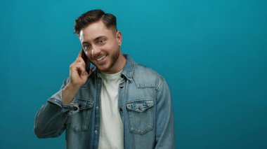 Sakallı genç adam mavi bir duvara karşı akıllı bir telefonla konuşurken gülümsüyor, kot ceket giyiyor, rahat bir şekilde giyiniyor ve izole bir şekilde iletişim kuruyor..