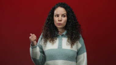 Kıvırcık saçlı kadın, kırmızı arka plana karşı kuşkulu bir ifade takınarak izole stüdyo ortamında şüphe ve belirsizliği işaret ediyor..