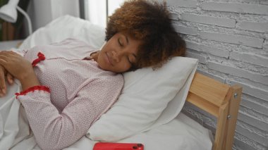 Kıvırcık saçlı kadın rahat bir yatakta uyur, pijama giyer, parlak bir yatak odasında..