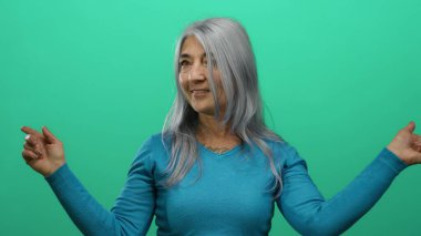 Gri saçlı, mavi kazaklı bir kadın canlı yeşil arka plana karşı yan yan bakıyor. Yön ve pozitifliği neşeli bir ifadeyle aktarıyor..