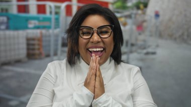 Bir inşaat alanında gülümseyen, iri yarı, gözlüklü, beyaz gömlekli, sanayileşmiş bir ortama güveni ve güveni olan genç, Latin bir kadın..