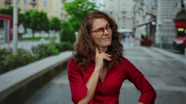 Kırmızı tişörtlü, kıvırcık saçlı ve gözlüklü bir kadın. Arka planda bulanık binalar ve yeşil ağaçlarla duygularını ifade ediyor..