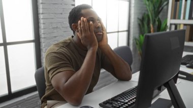 Genç adam kapalı bir masada oturmuş modern ofis ortamında düşünceli bir ifadeyle bilgisayara bakıyor. Doğal ışık ve kapalı alan bitkiler.