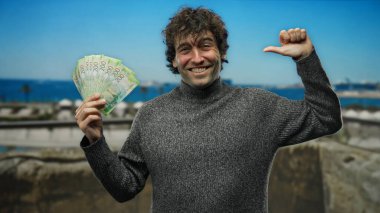 İspanyol adam, elinde Rus rublesi tutarak güneşli bir deniz kenarına işaret ediyor. Arka planda dalgalar yavaşça sahile vuruyor..