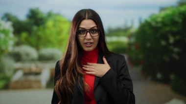 Kırmızı balıkçı yaka kazaklı ve gözlüklü siyah ceketli kadın yemyeşil parkta mahkumiyetle konuşuyor..