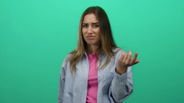 Genç bir kadın yeşil izole bir duvara karşı parmaklarıyla bir jest yapıyor, tetikte ve odaklanmış, günlük kıyafetler giyiyor ve sarı bir saç stili sergiliyor.