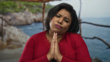Kırmızılı kadın sakin sahilde derin bir ifadeyle dua ediyor. Açık deniz sahnesinde doğanın, denizin ve iç huzurun uyumunu gösteriyor..