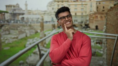 Gözlüklü genç adam şehirdeki antik Roma harabelerinde düşünceli bir şekilde duruyor, bulutlu bir gökyüzünün altında canlı kırmızı bir kazakla cazibe ve çekicilik yayıyor..