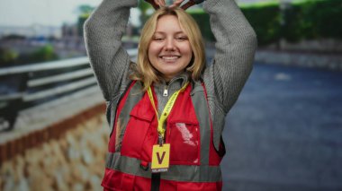 Şehir caddesinde kalp sembolü yapan, kırmızı yelek giyen ve açık havada gülümseyen sarı saçlı gönüllü bir kadın..