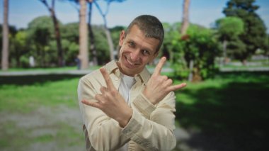 Kafkasyalı bir adam güneşli bir parkta, açık mavi gökyüzü altında yemyeşil yeşil bir ortamda, coşkuyla el kol hareketi yapar..