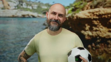 Güneşli deniz kıyısında futbol topu tutan sakallı kel bir adam. Arka planda kayalık bir sahil, açık hava karizması ve atletik ruhu somutlaştırıyor..