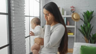 Modern bir apartman dairesinde bebeğini tutan kadın pencereden dışarı bakıyor. Evin içinde sevgi ve aile dinamiklerini tasvir ediyor. Şık dekorlar ve yeşil bitkilerle..