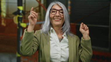 İnşaat alanında gülümseyen, gözlüklü, resmi kıyafetli, kıdemli, kır saçlı kadın şehir ortamında profesyonel bir ilişki ve mutluluğu öneriyor..