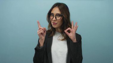 Gözlüklü İspanyol bir kadın. Mavi arka planda izole edilmiş bir tabela. Yüzünde anlamlı bir jest olan basit bir kıyafete profesyonel güvenini gösteriyor..