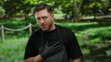 Sakallı, eldivenli ve çizgili önlüklü bir genç parkta gülümsüyor..