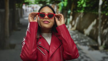 Kırmızı ceketli ve güneş gözlüklü bir kadın güneşli bir sokakta kendine güvenen bir stil sergiliyor..
