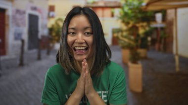 Genç bir kadın şehir merkezinde yeşil bir tişört giyerek alkışlıyor. Şehir ortamında neşeli ve canlı bir anı yakalıyor..