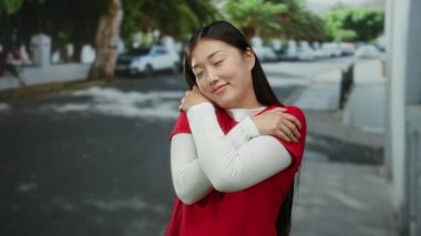 Kırmızı tişörtlü kadın sakin bir şehir caddesinde dikiliyor gözleri kapalı, ağaçlar ve park halindeki arabalarla şehir ortamında huzurlu bir ifade sergiliyor..