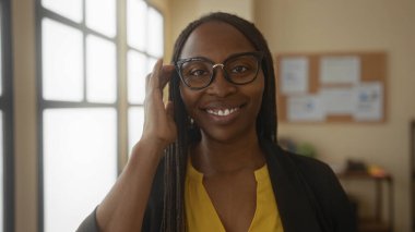 Ofis ortamında gözlüklü gülümseyen, parlak camlı, sarı gömlekli, kıvırcık saçlı, kendine güveni ve profesyonelliği olan bir kadın..