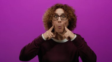 Canlı pembe bir arka plana karşı komik bir ifade sergileyen kadın, kıvırcık saçlı, gözlüklü ve günlük kıyafetleriyle mizah ve kişilik sergiliyor..