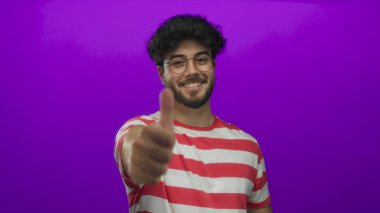 Sakallı, çizgili tişörtlü ve gözlüklü genç adam canlı mor arka planda parmaklarıyla güvenle gülümsüyor..