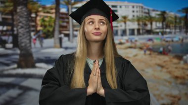 Mezuniyet üniformalı genç bir kadın, arka planda deniz ve kumsal olan güneşli bir deniz kenarında dua ederken poz veriyor..