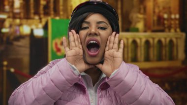 Kilisenin içinde kask ve pembe ceket giyen genç bir kadın, enerjik bir şekilde el kol hareketi yaparak, kapalı bir yerde bulanık bir dini zemine karşı canlı bir ifade yakalıyor..