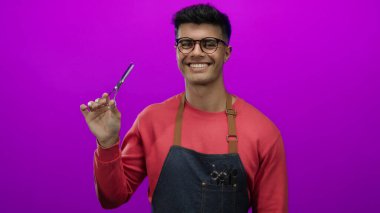 Genç, İspanyol asıllı bir adam parlak pembe bir arka plana karşı kuaför makası tutuyor. Gözlüklü, kırmızı kazaklı ve kot önlüklü, modern berber tarzı giyiniyor..