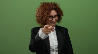 Gözlüklü bir kadın titiz bir jest yapıyor, düz yeşil bir arka plana karşı duruyor, resmi bir takım elbise giyiyor, farkındalık ve ciddiyet duygusunu gösteriyor..