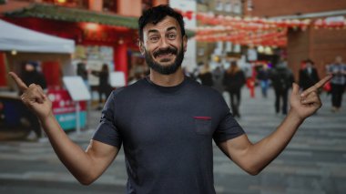 Sakallı, gülümseyen ve işaret eden bir adam hareketli bir şehir caddesinde dikilirken arka planda neşeli insanlar ve şenlikli dekorasyonlarla.
