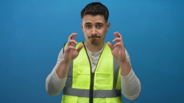 Yansıtıcı yelek giyen genç İspanyol adam umut ya da batıl inanç gösteren izole edilmiş mavi arka plan duvarına karşı şans diliyor..