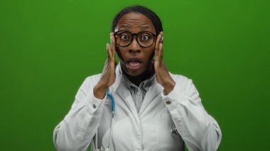 Gözlüklü ve steteskop takan doktor, yeşil duvara karşı duruyor. Yüzü üzerinde çeşitli duygu ve hareketleri inceliyor. Klinik ortamında odaklanmış ve yoğunlaşmış görünüyor..