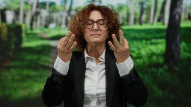 Parkta nefes egzersizi yapan İspanyol kadın gözlük ve takım elbise takıyor, açık havada yemyeşil ağaçlar ve çimlerle çevrili..