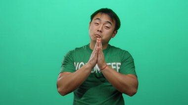 Yeşil gömlekli genç adam yeşil arka planda el ele poz veriyor, yakışıklı ve düşünceli bir ifade sergiliyor, barış ve tefekkür öneriyor..