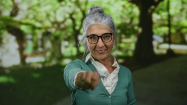 Açık hava parkında gülen gri saçlı yaşlı bir kadın. Etrafı yemyeşil yeşillik ve güneş ışığıyla çevrili..