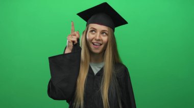 Mezuniyet üniformalı genç beyaz kadın yeşil arka planda kutlama yapıyor, neşeli bir ifadeyle, başarıyı ve başarıyı sembolize ediyor..