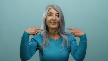 Gri saçlı yaşlı bir kadın kendini mavi bir arka plan duvarına doğru gösteriyor..
