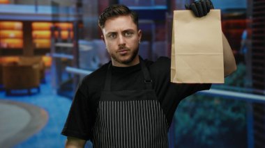 Önlük takan sakallı genç adam otel ortamında kese kâğıdı tutuyor. Zarif bir yemek ortamında yemek servisi yapıyor..