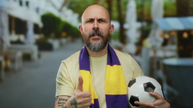 Sakallı bir adam futbol topuyla baş parmağını kaldırıyor, atkı takıyor, açık hava kafeleri olan canlı bir şehir sokağında duruyor, coşkuyu ve takım ruhunu yansıtıyor..