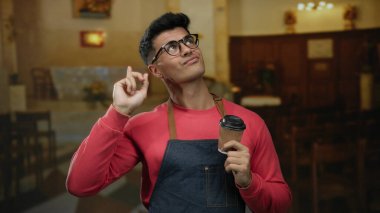 Kahve fincanlı genç adam kırmızı kazak ve önlük giyiyor, otel ortamında gülümsüyor, özgüven ve cazibe yayıyor, içeri davet edici bir hava katıyor..