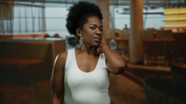 Beyaz elbiseli kadın boynuna dokunuyor, modern restoran içlerine karşı yumuşak ışıklandırma, rahatlık ve tarz taşıyor..